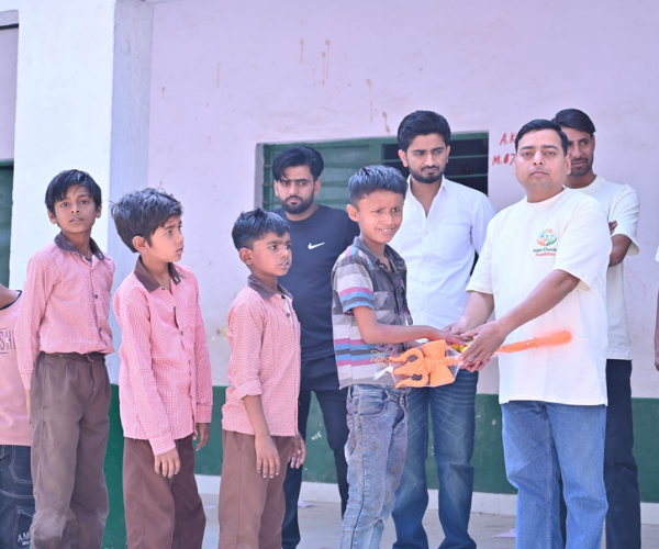 Foundation volunteer handing a saffron ceremonial trishul gift to a boy while schoolchildren in uniform watch